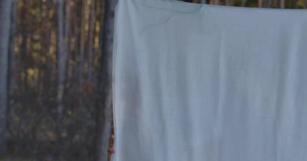 Joven chica positiva caucásica escondida detrás de ropa blanca en el bosque. Hermosa chica disfrutando de calma día de verano al aire libre . — Vídeo de stock