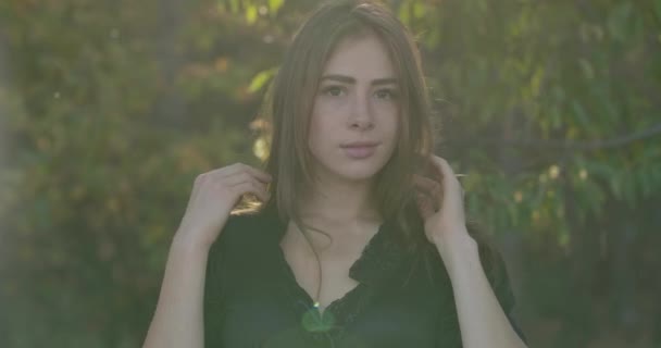 Verschwommenes Porträt einer netten kaukasischen brünetten Frau mit braunen Augen im schwarzen Kleid, die zwischen Bäumen im Wald steht. Charmantes Mädchen, das in die Kamera schaut und lächelt. — Stockvideo