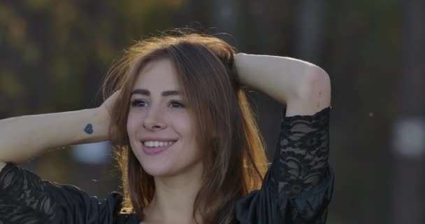 Close-up retrato de uma mulher morena branca sorridente agradável com olhos castanhos em vestido preto de pé entre as árvores na floresta. Menina encantadora desfrutando de noite tranquila ao ar livre . — Vídeo de Stock