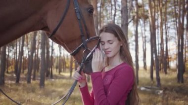 Pembe elbiseli, sonbahar ormanında at okşayan ve gülümseyen beyaz bir kızın yakın çekimi. Güzel esmer at binicisi sevimli hayvanıyla dışarıda dinleniyor..