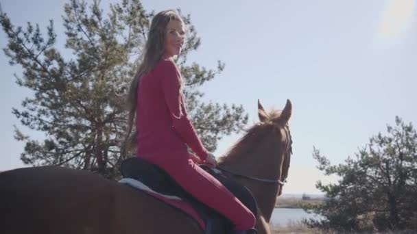 Pohled zezadu na usmívající se bělošku sedící na koni na slunci a ujíždějící pryč. Roztomilá brunetka dívka v růžovém oblečení odpočívá se svým domácím mazlíčkem venku. — Stock video