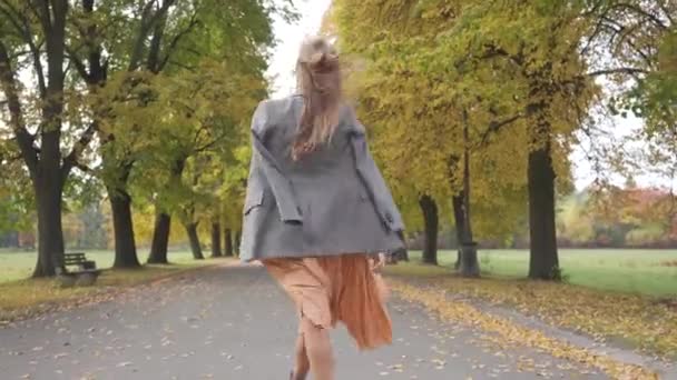 На задньому плані - струнка кавказька жінка у викарбуваній куртці та гірчичній сукні, що йде в осінньому парку. Дівчинка прогулювалась по дорозі і обернулася, щоб зазирнути в камеру.. — стокове відео