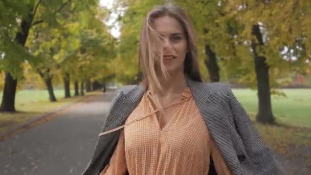 Close-up of a slim Caucasian woman in checkered jacket and mustard dress turning around and strolling along the road. Beautiful brunette girl walking in the autumn park. — Stock Video