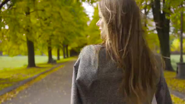 Вид сзади на улыбающуюся белую женщину в клетчатой куртке, идущую по дороге. Юная брюнетка прогуливается в осеннем парке . — стоковое видео