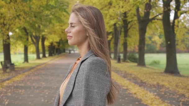 Menina morena caucasiana confiante olhando ao redor e andando ao longo da estrada. Mulher sorridente agradável em jaqueta quadriculada passeando no parque de outono . — Vídeo de Stock