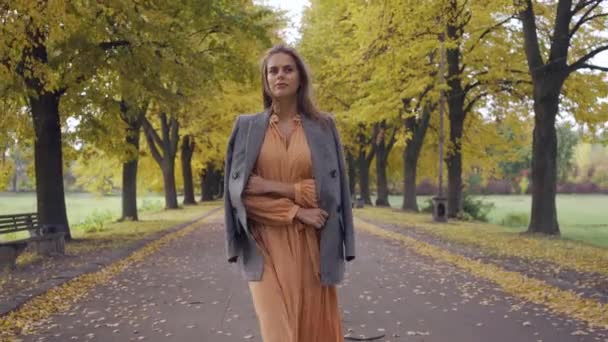 Portrait of a confident Caucasian girl in mustard dress and checkered jacket walking along the road. Pretty brunette woman strolling in the autumn park on weekends. — ストック動画