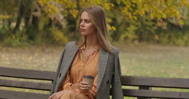 Wanita Kaukasia cantik duduk di bangku cadangan di taman musim gugur dengan secangkir kopi. Gadis yang menarik mengguncang rambut coklatnya yang panjang dan berpaling. Bioskop 4k rekaman ProRes HQ . — Stok Video