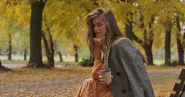 Vista lateral de la encantadora mujer caucásica con el pelo castaño largo y los ojos verdes sentados en el banco en el parque de otoño. Chica atractiva sosteniendo una taza de café y sonriendo. Cinema 4k metraje ProRes HQ . — Vídeos de Stock