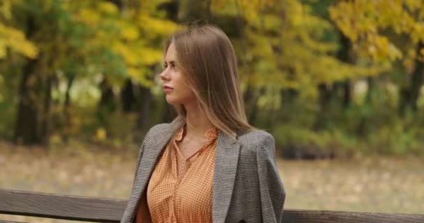 Profile portrait of an elegant Caucasian woman sitting on the bench in the park and smiling. Charming girl in checkered jacket and mustard dress spending time outdoors. Cinema 4k footage ProRes HQ. — ストック動画