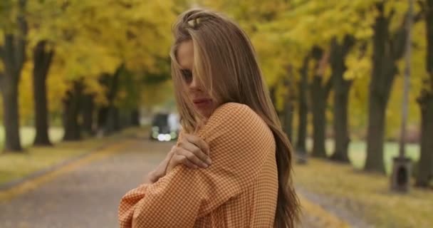 Vista lateral de la chica caucásica congelada con el pelo castaño y sonrisa agradable de pie en la carretera en el parque de otoño. Mujer bonita en vestido de mostaza con patas de cuervo estampado metiendo el pelo. Cinema 4k metraje ProRes HQ . — Vídeos de Stock