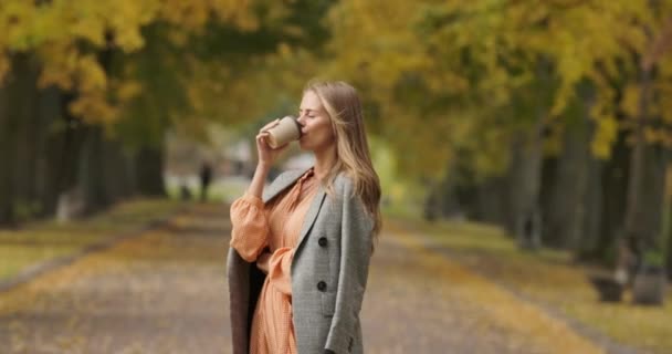 Elegante mujer caucásica con cabello castaño largo vestida con chaqueta a cuadros bebiendo café en el parque de otoño. Hermosa mujer girando a la cámara y sonriendo. Cinema 4k metraje ProRes HQ . — Vídeos de Stock