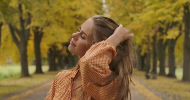 Close-up portrait of cute Caucasian girl standing in autumn park and sighing. Attractive woman in mustard dress with tweed print holding long brown hair in wind. Cinema 4k footage ProRes HQ. — ストック動画