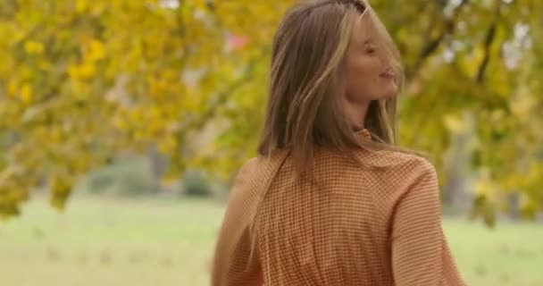 Smiling Caucasian girl with long brown hair and green eyes spinning in the autumn park. Elegant woman in mustard dress with crows feet print having fun in the autumn park. Cinema 4k footage ProRes HQ — ストック動画
