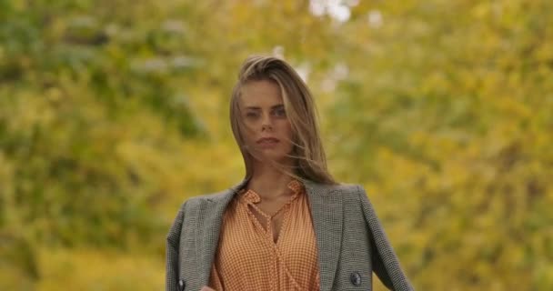 Young Caucasian girl with green eyes standing on the background of autumn leaves and looking at camera. Woman in mustard dress and checkered jacket holds long hair in wind Cinema 4k footage ProRes HQ — ストック動画