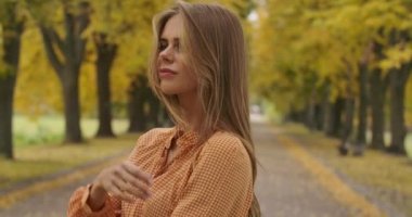 Sonbahar parkında yolda duran kendine güvenen beyaz bir kadının portresi. Hardal elbiseli, saçları kulağının arkasında kıvrılıp gülümseyen hoş bir kız. Sinema 4k görüntüleri Prores Hq.