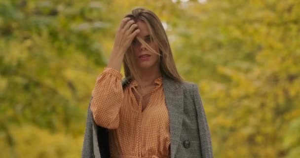 Young Caucasian woman with green eyes standing on the background of leaves and looking at camera. Pretty woman in mustard dress and checkered jacket touching her long hair. Cinema 4k footage ProRes HQ — Stock Video