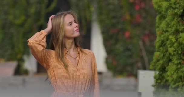 Attractive Caucasian woman in mustard dress touching her head with hands and smiling. Cute girl standing on the background of house covered with green and red bushes. Cinema 4k footage ProRes HQ. — Stock Video