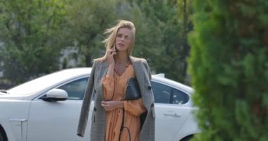 Gülümseyen beyaz kadın arabanın yanında duruyor ve cep telefonuyla konuşuyor. Uzun kahverengi saçlı, kendine güvenen iş kadını hoş sohbetler ediyor. Sinema 4k görüntüleri Prores Hq.