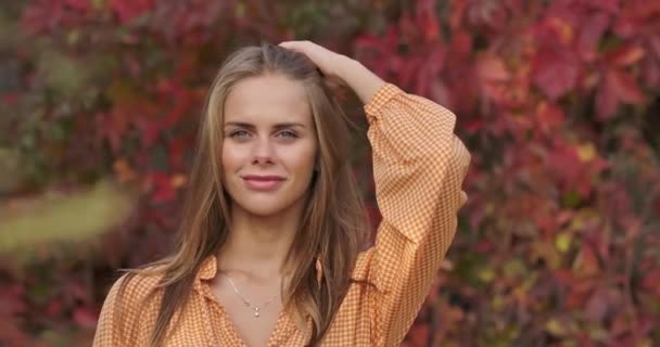 Primo piano di ragazza sorridente in piedi sullo sfondo di foglie rosse e toccare i capelli castani. Affascinante donna sicura di sé in abito di senape in posa alla fotocamera nel parco autunnale. Cinema 4k riprese ProRes HQ . — Video Stock