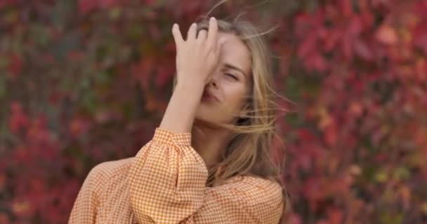 Close-up of confident lady in mustard dress standing on the background of red leaves and holding her long hair in wind. Charming woman posing at camera in autumn park. Cinema 4k footage ProRes HQ. — ストック動画