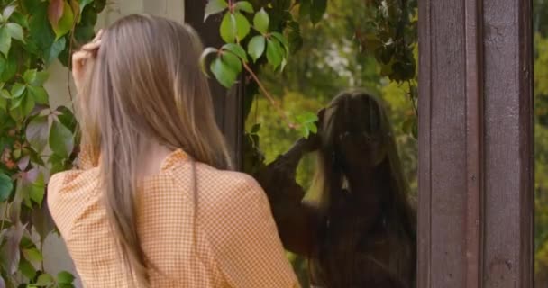 Back view of a young brunette Caucasian woman in mustard dress looking at the glass doors. Slim girl fixing her long hair using door as a mirror. Cinema 4k footage ProRes HQ. — ストック動画