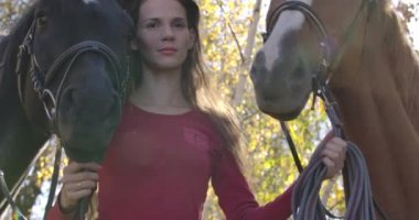İki atın dizginlerini tutan, derisi sorunlu, üzgün, beyaz bir kadın. Güneşli sonbahar ormanlarında hipoterapi gören hasta bir kadın. Sinema 4k görüntüleri Prores Hq.