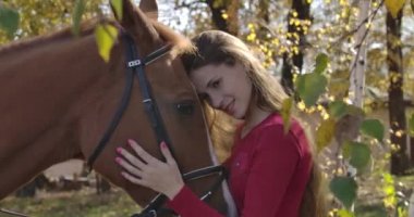 Sonbahar ormanında at suratlı gülümseyen beyaz bir kızın yakın çekimi. Güzel esmer at binicisi güzel hayvanıyla dışarıda dinleniyor. Sinema 4k görüntüleri Prores Hq.