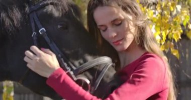 Sonbahar ormanında at suratını okşayan ve gülümseyen beyaz bir kızın yakın plan portresi. Güzel esmer at binicisi güzel hayvanıyla dışarıda dinleniyor. Sinema 4k görüntüleri Prores Hq.