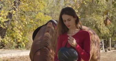 Profesyonel beyaz kadın jokey ata binme kaskı takıyor. Sonbahar ormanında güneş ışığında kahverengi atıyla duran genç bir kızın portresi. Sinema 4k görüntüleri Prores Hq.