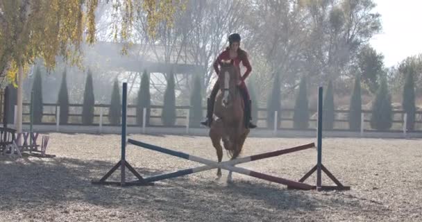 Young Caucasian female equestrian in helmet jumps the barrier on graceful brown horse and riding away from shot. Professional female equestrian training in corral. Cinema 4k footage ProRes HQ. — Stock Video