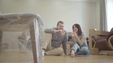 Genç, beyaz bir aile oturma odasında yerde fincanlarla oturuyor ve yeni mobilyalarla konuşuyor. Mutlu çift yeni evlerine taşınıyor. Yeni bir hayatın başlangıcı.