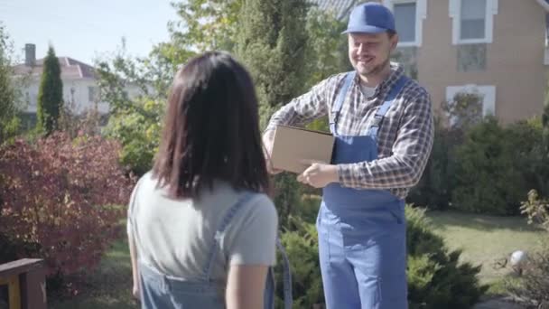 Lächelnder kaukasischer Kurier in blauer Arbeitskleidung, der der Kundin einen Karton überreicht und darauf wartet, dass sie das Paket kontrolliert. Lieferservice nach Hause, Online-Shopping. — Stockvideo