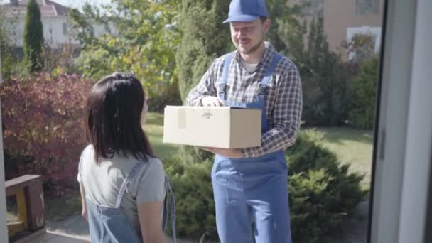 Positivo mensajero masculino caucásico en ropa de trabajo azul dando caja a la cliente femenina y esperando a que firme la hoja de ruta. Servicio de entrega a domicilio, compras en línea . — Vídeos de Stock