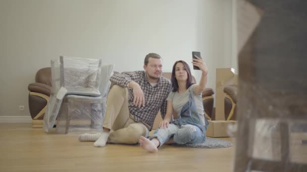 Glad vit par tar selfie som sitter på golvet i vardagsrummet med nya möbler. Ett leende par flyttar in i sitt nya hus. Början på nytt liv. — Stockvideo