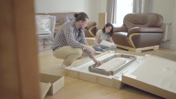 Young positive Caucasian family unpacking parcel in the living room. Happy couple furnishing their new house. Start of future life. — Stock Video