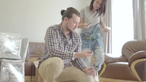 Jonge blanke echtpaar monteert nieuw meubilair in de woonkamer. Gelukkige familie die hun nieuwe huis inrichten. Begin van het toekomstige leven. We komen eraan.. — Stockvideo