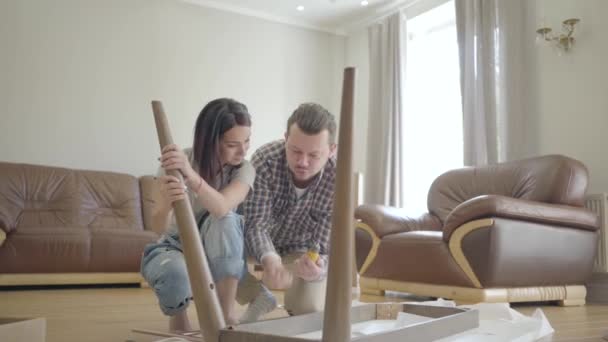 Portrait of a young Caucasian family assembling table in the living room. Positive family furnishing their new house. Moving in. — Stock Video