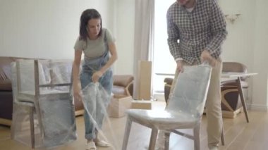 Genç, güzel, beyaz bir çift oturma odasında yeni bir sandalye açıyor. Mutlu aile eve taşınıyor. Yeni bir hayatın başlangıcı.