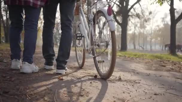 Közelkép két pár felnőtt lábról tornacipőben, ahogy biciklivel sétálnak a park sikátorában. Család vagy barátok töltik az őszi estét a szabadban. Egészséges életmód koncepció. — Stock videók