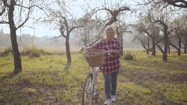 Leende mogen vit kvinna i casual kläder promenader i dimmig park med cykel. Vacker europeisk pensionerad dam tillbringar höstkvällen utomhus. Glad senior på semester. — Stockvideo