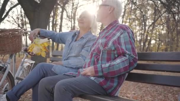 Glad vit pensionerad par sitter på bänken i solljus och pratar. Gammal europeisk familj spenderar solig kväll utomhus. Åldrande tillsammans, evig kärlek koncept. — Stockvideo