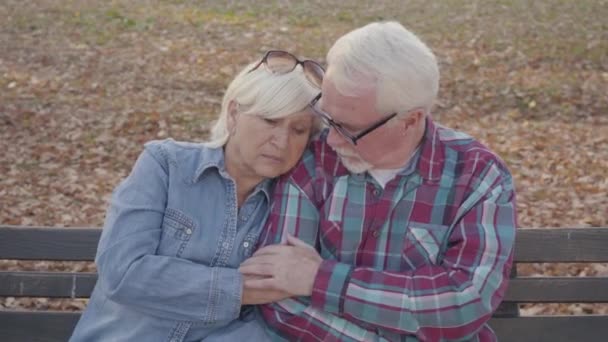 Retrato de una pareja jubilada europea sentada en el banquillo y hablando. Marido maduro calmando a su hermosa esposa mayor. Envejecer juntos, concepto de amor eterno . — Vídeos de Stock