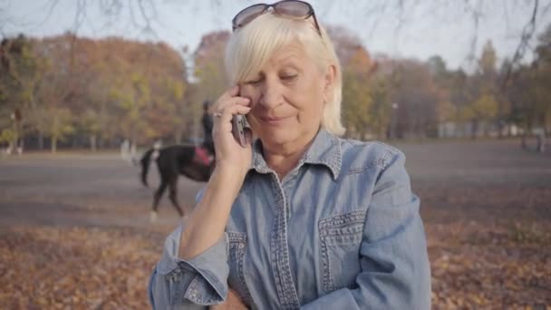 Ritratto di donna matura europea in piedi nel parco autunnale e che parla con lo smartphone. Donna caucasica anziana positiva che riposa all'aperto. Bella pensionato femminile godendo di tempo libero . — Video Stock