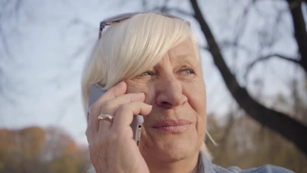 Közelkép egy érett európai nőről, aki az őszi parkban áll és okostelefonon beszél. Pozitív idős fehér hölgy pihen a szabadban. Gyönyörű női nyugdíjas élvezi a szabadidőt. — Stock videók