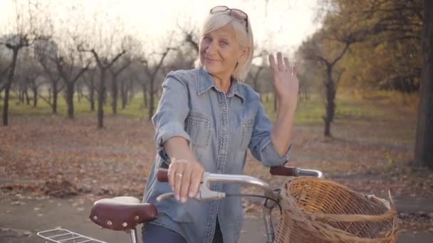 Femme blanche mature positive en vêtements décontractés debout avec vélo et agitant à la caméra. Belle retraitée européenne passant la soirée d'automne à l'extérieur. Happy senior en vacances . — Video