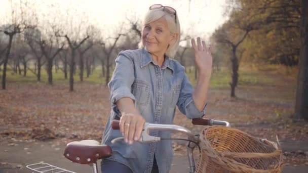 Mujer caucásica madura positiva en ropa casual de pie con bicicleta y saludando a la cámara. Hermosa señora jubilada europea pasar la noche de otoño al aire libre. Feliz senior en vacaciones . — Vídeos de Stock