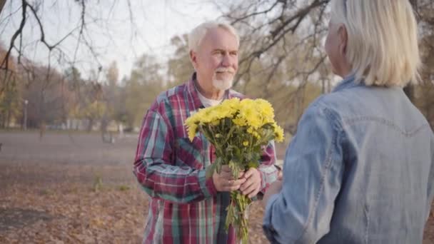 Homem maduro caucasiano feliz dando bouquet crisântemo para sua adorável esposa e ajoelhado. Família madura europeia positiva passando noite romântica no parque de outono. Envelhecimento juntos, amor eterno . — Vídeo de Stock