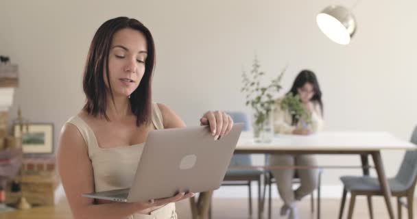 Porträt eines hübschen kaukasischen Mädchens, das den Laptop schließt und zu tanzen beginnt. ihre Freundin oder Schwester, die im Hintergrund am Tisch sitzt. positive Frauen, die sich zu Hause ausruhen. Kino 4k Filmmaterial prores hq. — Stockvideo