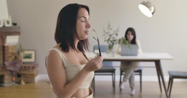 Joven chica caucásica mirando el fondo a su amiga o hermana sentada en la mesa, volviendo a la cámara y poniéndose gafas. Dos hermosas mujeres descansando en casa. Imágenes de Cinema4k ProRes HQ — Vídeo de stock