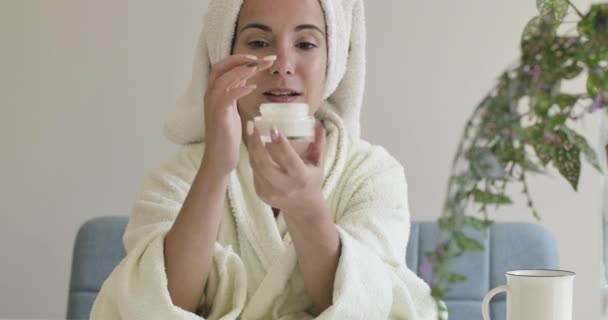 Portrait d'une jeune fille caucasienne en peignoir blanc et serviette de toilette appliquant de la crème visage. Belle femme essayant hydratant dans le salon de beauté ou à la maison. Cinéma 4k séquences ProRes HQ . — Video